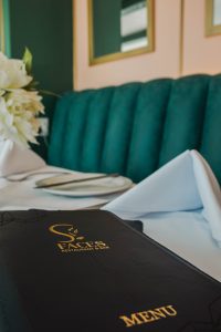 Close-up of a dark menu with gold lettering resting on a white-table setting beside a folded napkin and plate, with teal upholstery in the background.
