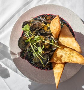 Bowl of steamed mussels with herbs and crumbled breadcrumbs, served with toasted garlic bread on a patterned plate