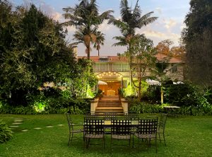 Outdoor garden seating in front of a warmly lit building entrance at dusk, with palm trees and lush greenery.