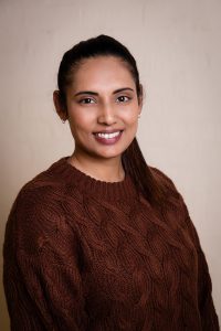 Portrait of a smiling woman with dark hair pulled back, wearing a brown cable-knit sweater against a beige background.