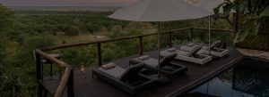 Wooden deck with four wicker lounge chairs, a large white umbrella, and towels, overlooking a green savannah at sunset—pool on the side.