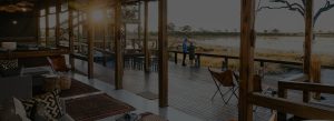 Wooden safari lodge lounge with exposed beams and a sunlit deck overlooking a water-filled plain at dusk, seating scattered inside and outside.
