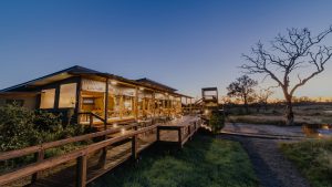 Wooden lodge with a glass-walled dining area on a raised deck, warm lights, at dusk in a dry savannah.