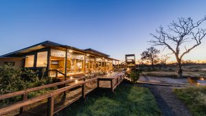 Wooden lodge with lit outdoor dining area along a boardwalk at sunset in a dry savanna with a bare-branched tree on the right